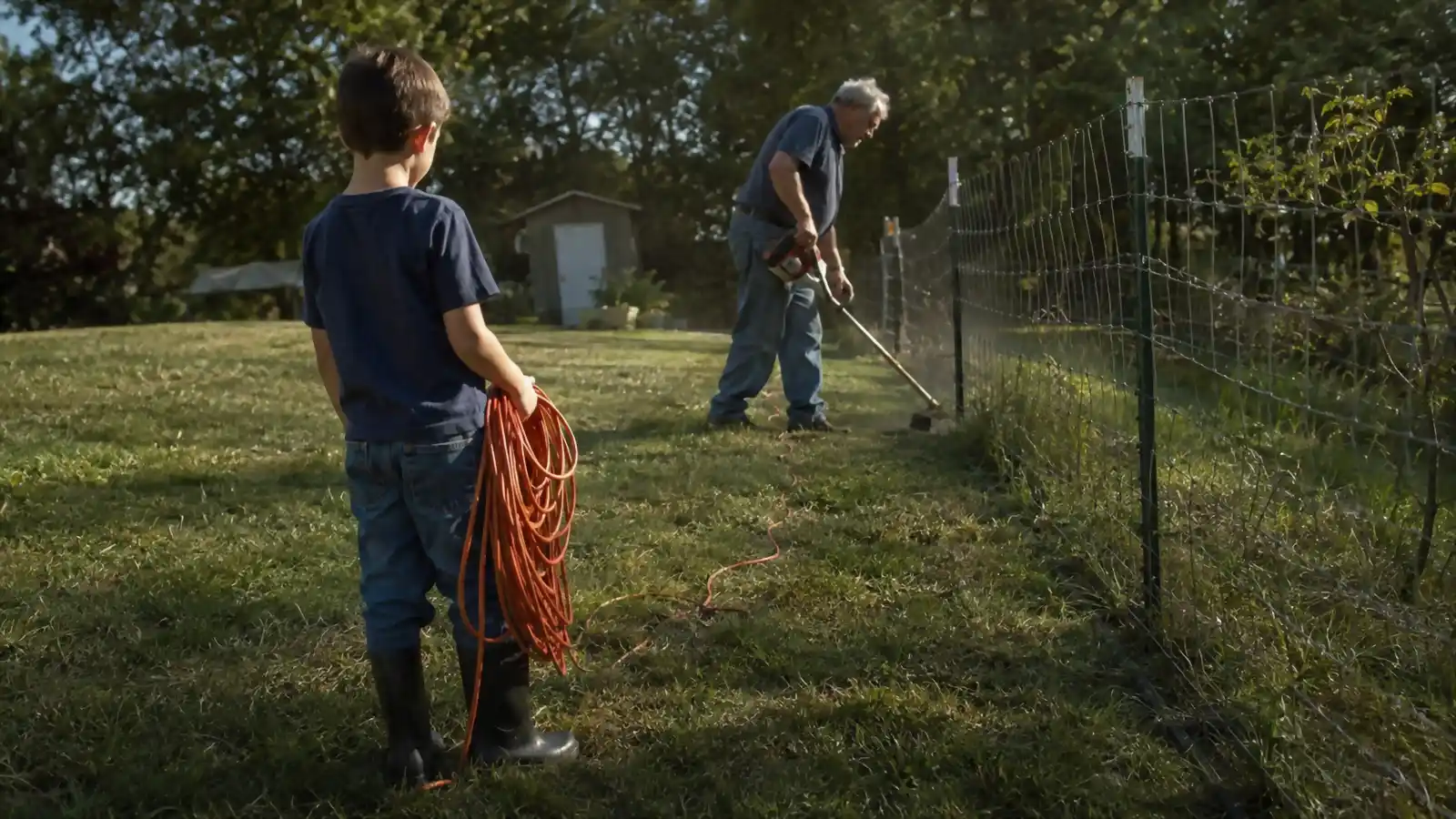 A boy standing near a fence line holding an extension cord while an older man trims grass nearby